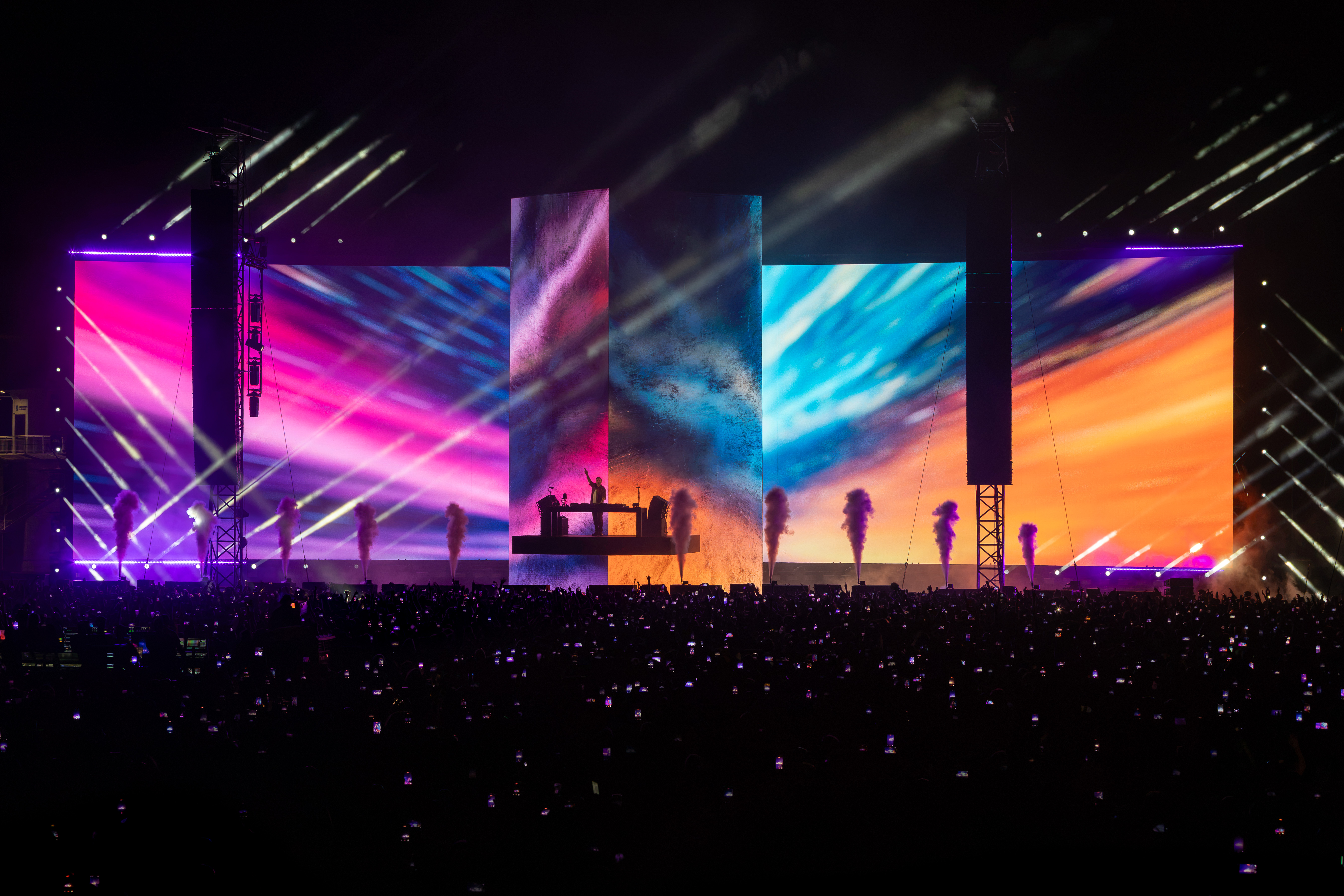 David Guetta's silhouette on the Monolith stage between two towering LED screens lit in pink, blue and orange, with a phone-lit crowd below at Parque Papa Francisco