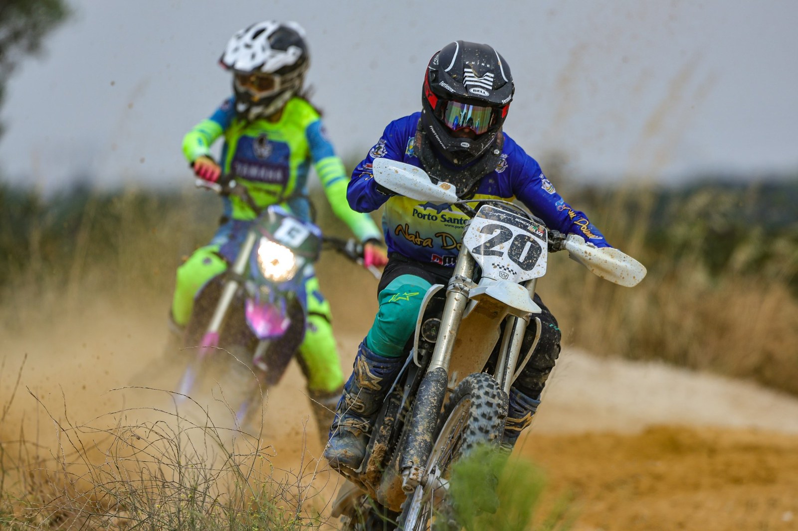 A rider in a blue and yellow Porto Santo jersey leads a competitor through a dusty section of track during the Fifty Party Motocross Experience in Pontes