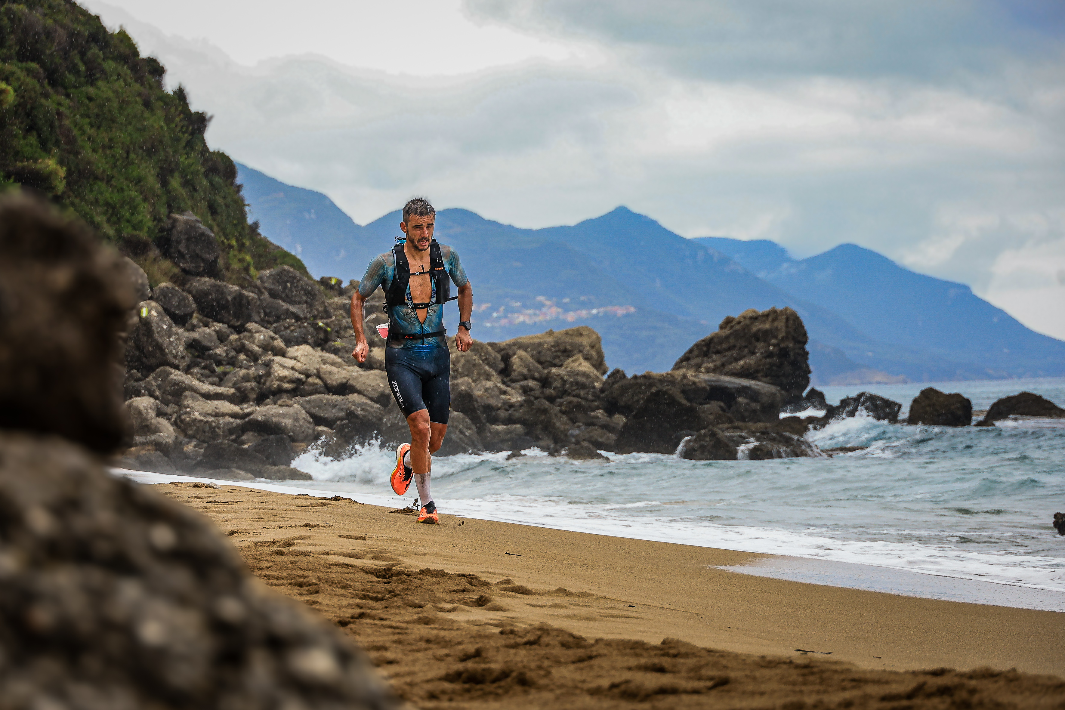 A competitor runs along a sandy shoreline beside rocks and breaking waves