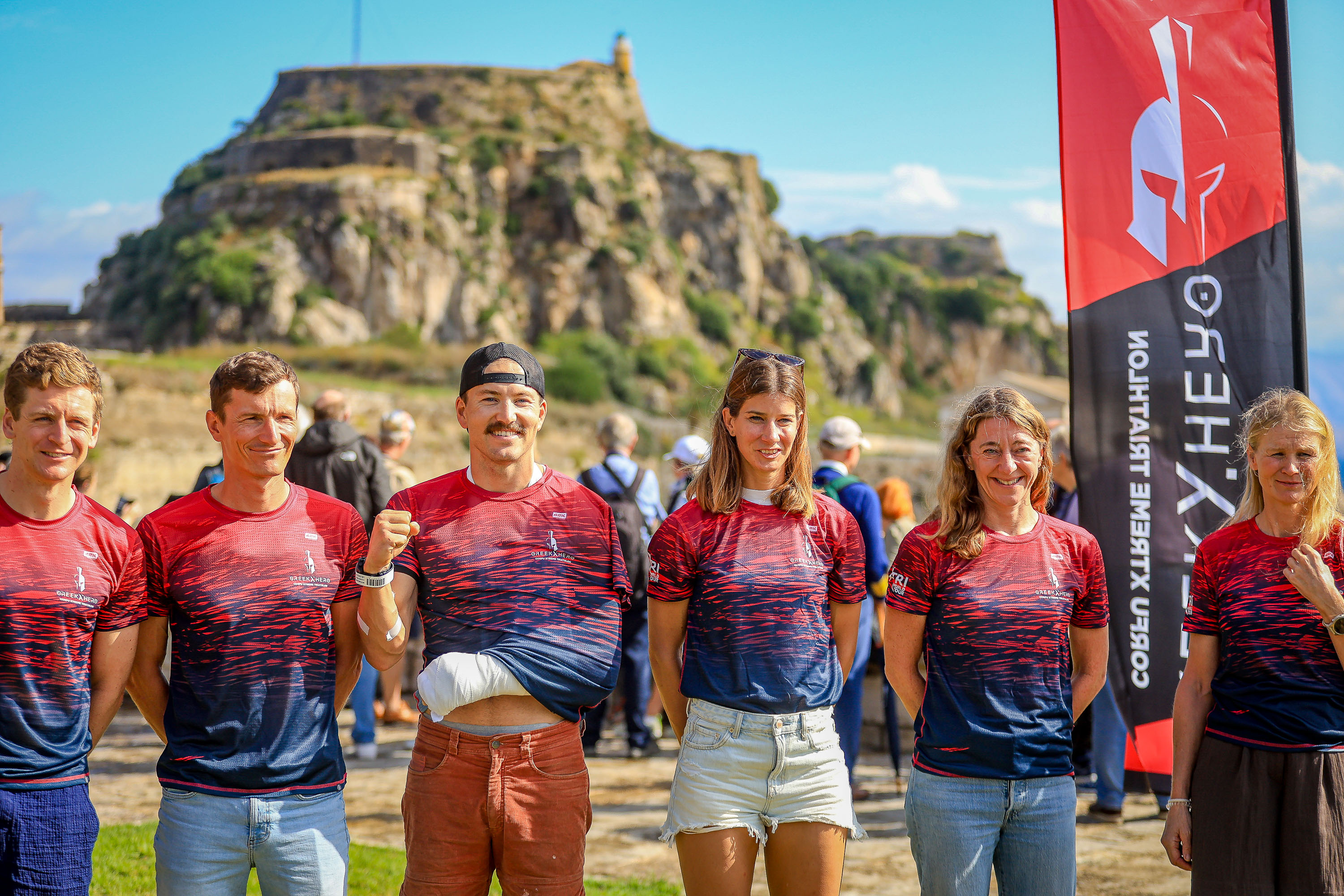 A group in Greek Hero event shirts poses beside a race flag