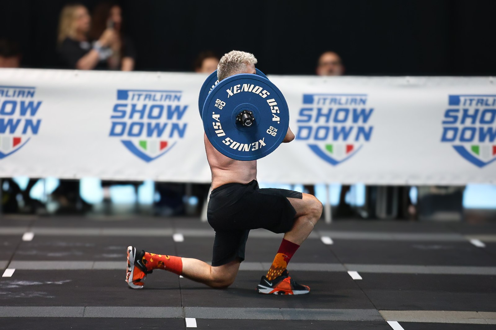 An athlete lunges overhead with a loaded barbell during Italian Showdown, back to camera, with an Italian Showdown banner behind him
