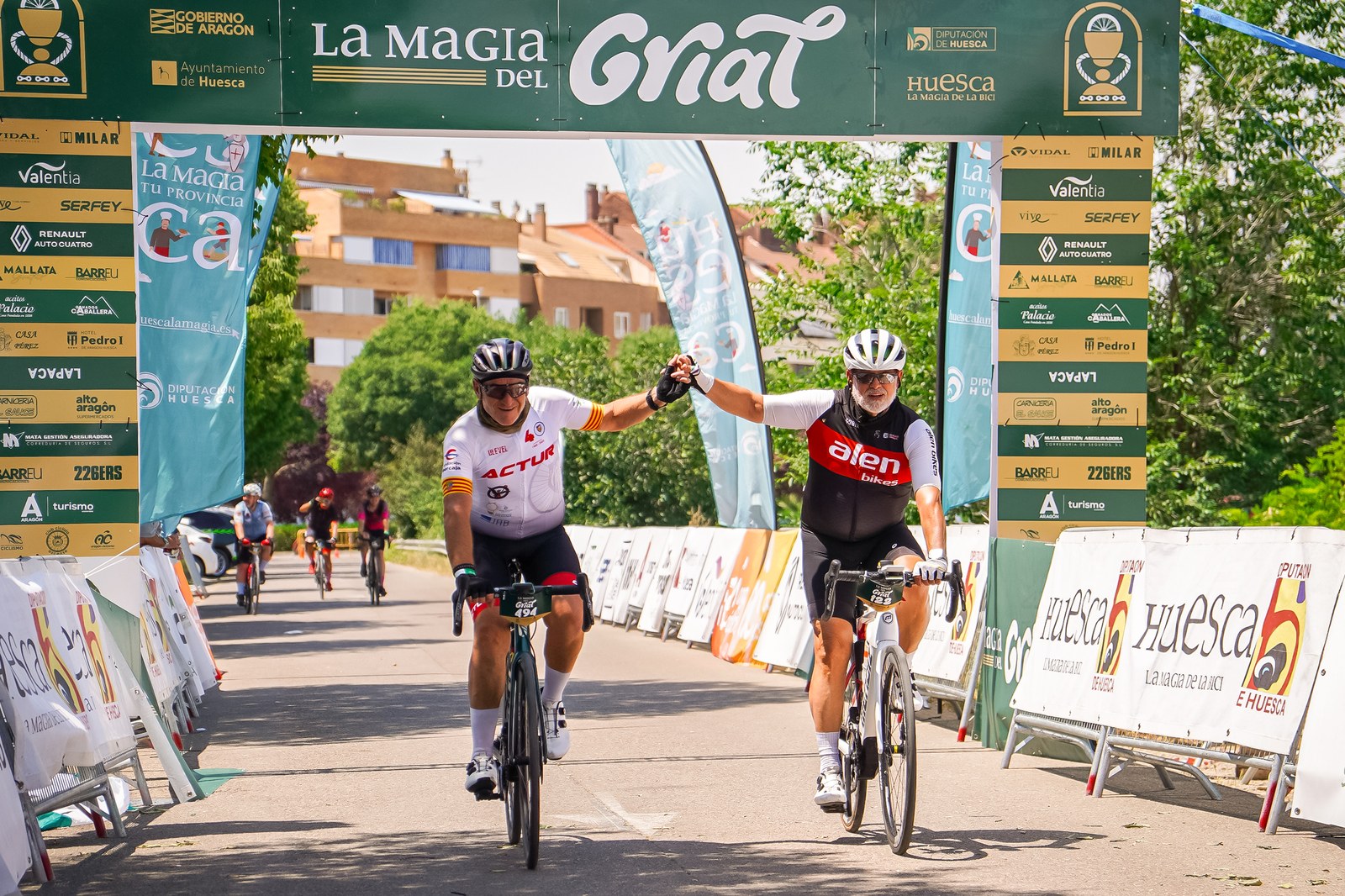 Two cyclists join hands as they cross the finish line together under the La Magia del Grial banner in Huesca