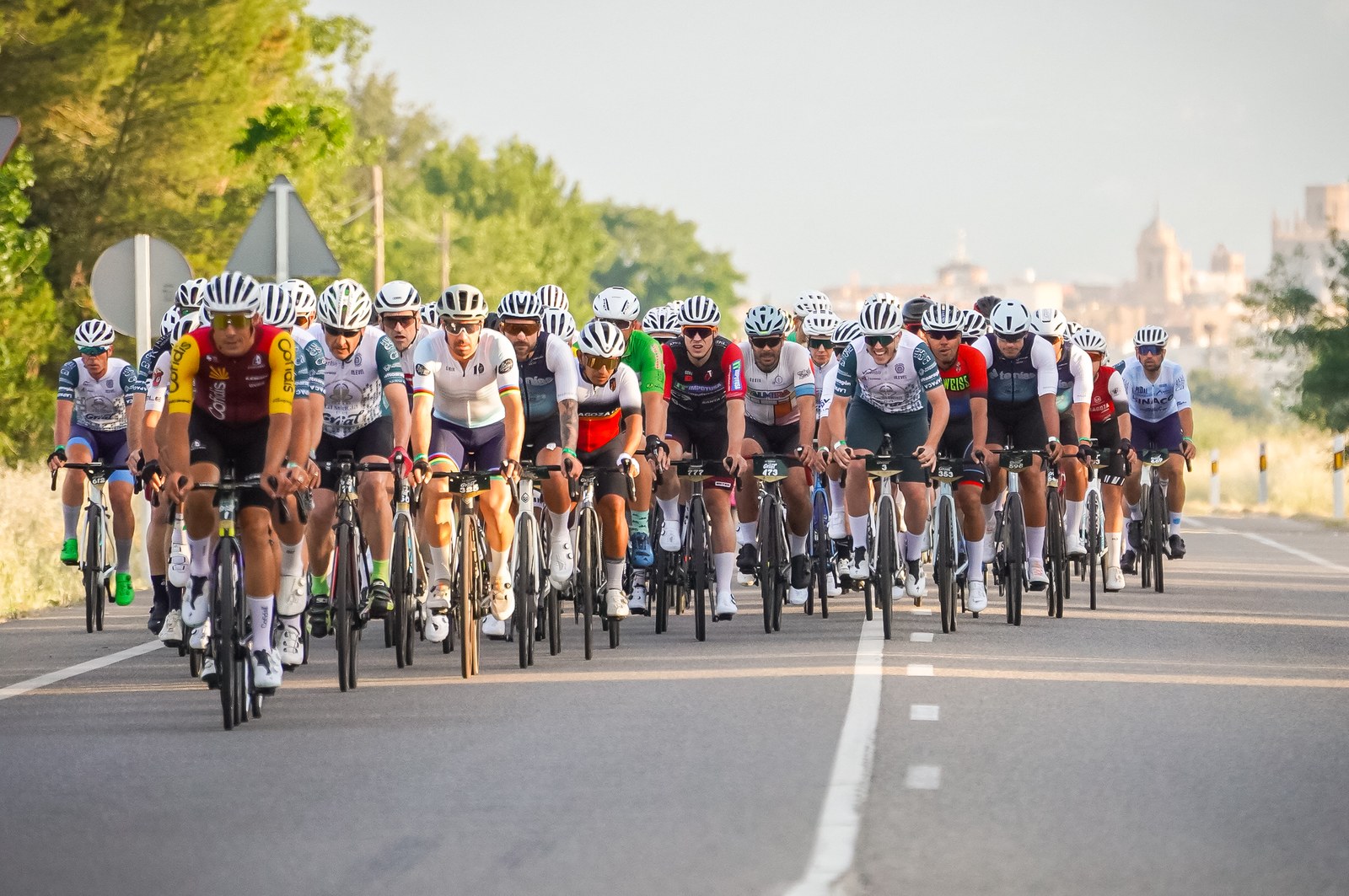 A large group of cyclists rides together on an open road during La Magia del Grial, with Huesca visible in the distance