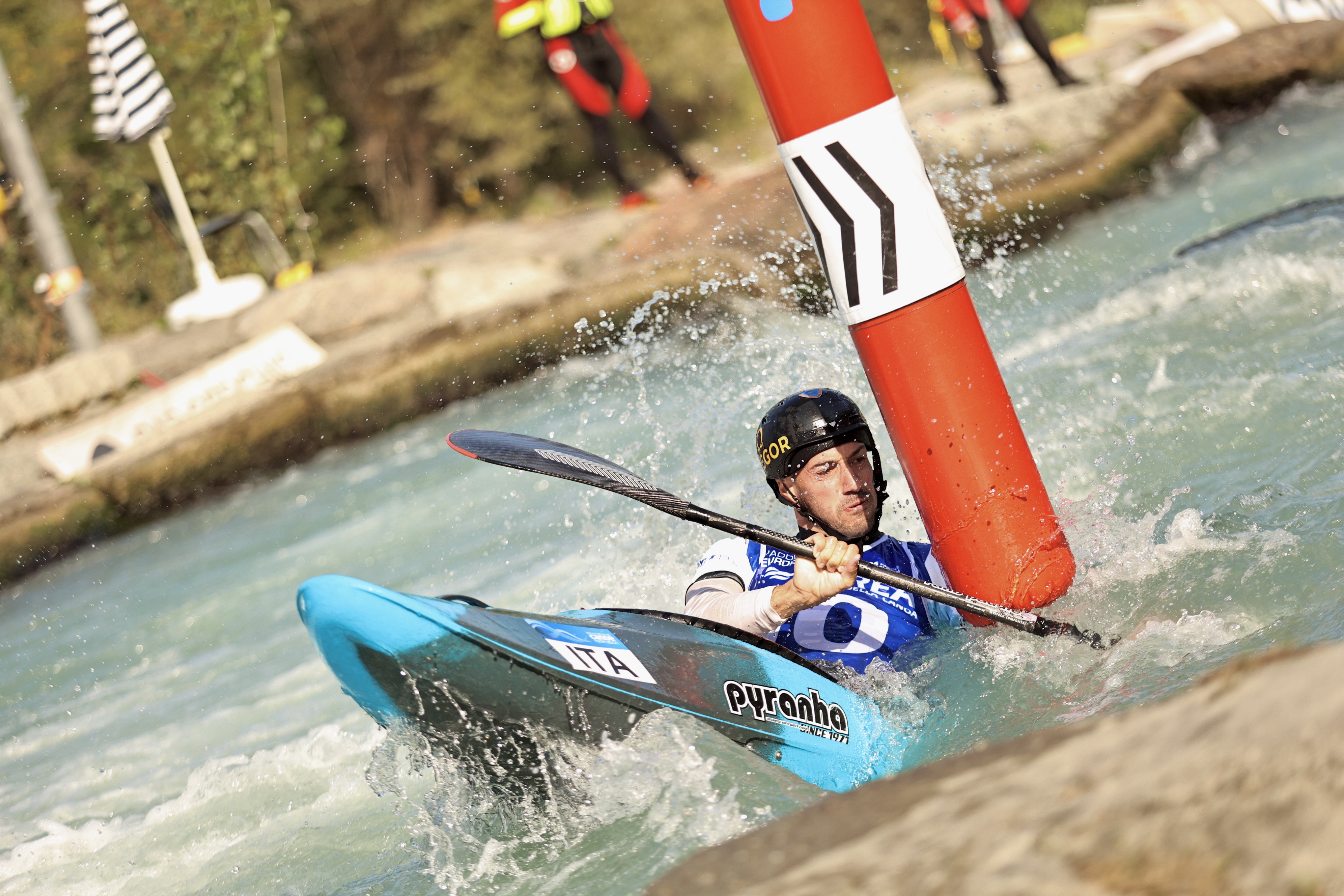 A canoe slalom athlete navigates the course beneath a red gate at the Ivrea Canoe Stadium