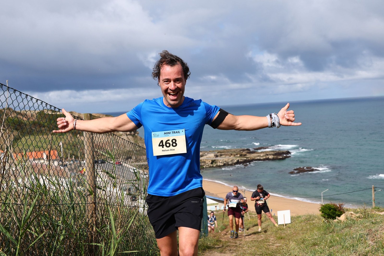 A runner celebrates with arms outstretched along the coastal trail at the Trail Rota dos Piratas