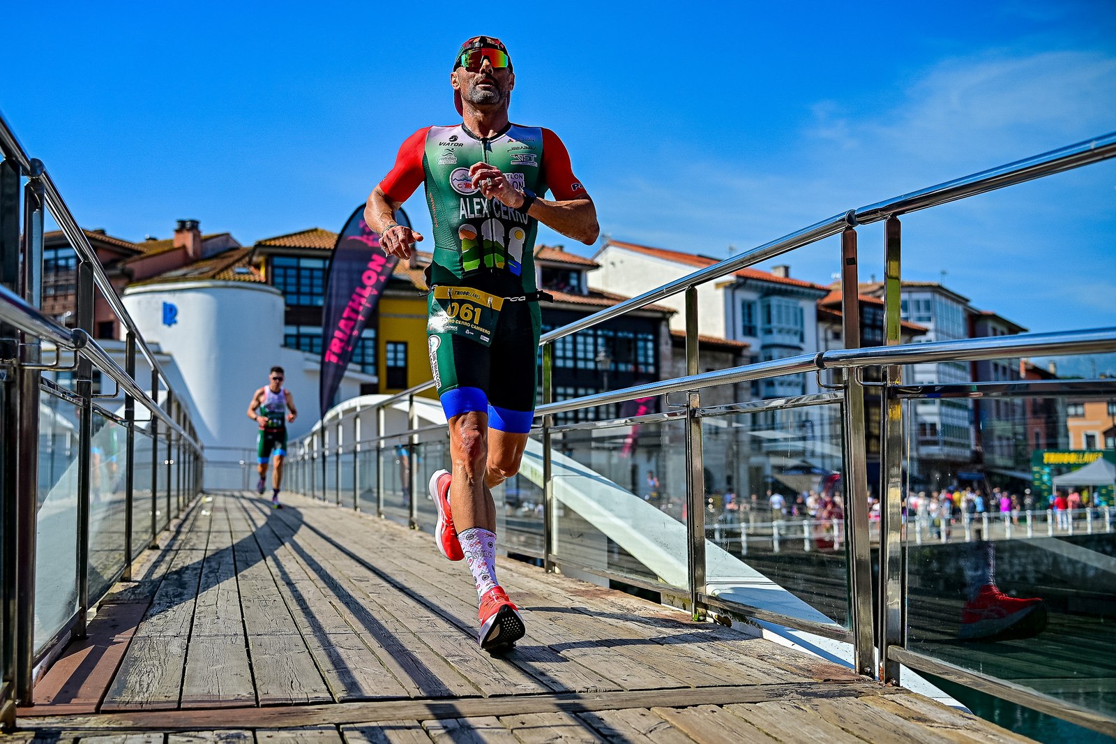 A runner crosses a footbridge over Llanes harbour during the Tri100Llanes run leg, with the town's coloured houses behind him