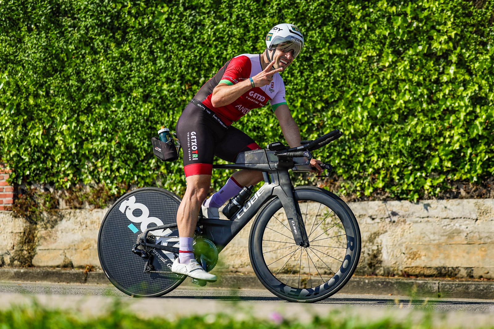 A cyclist on an aero triathlon bike smiles and gestures a peace sign during the Tri100Llanes bike leg