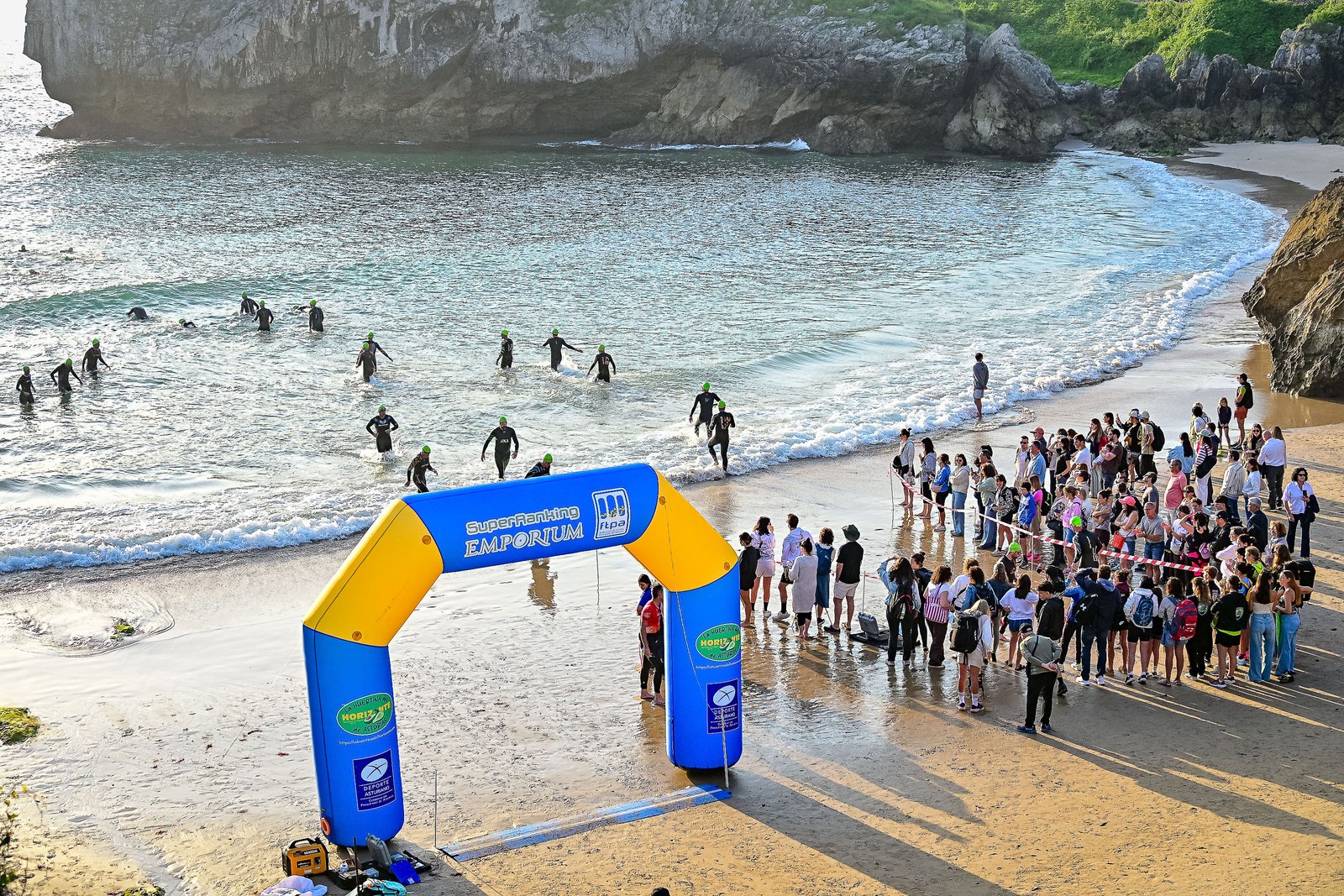 Aerial view of triathletes wading into the Cantabrian Sea at the start of the Tri100Llanes swim leg, with spectators lining the beach