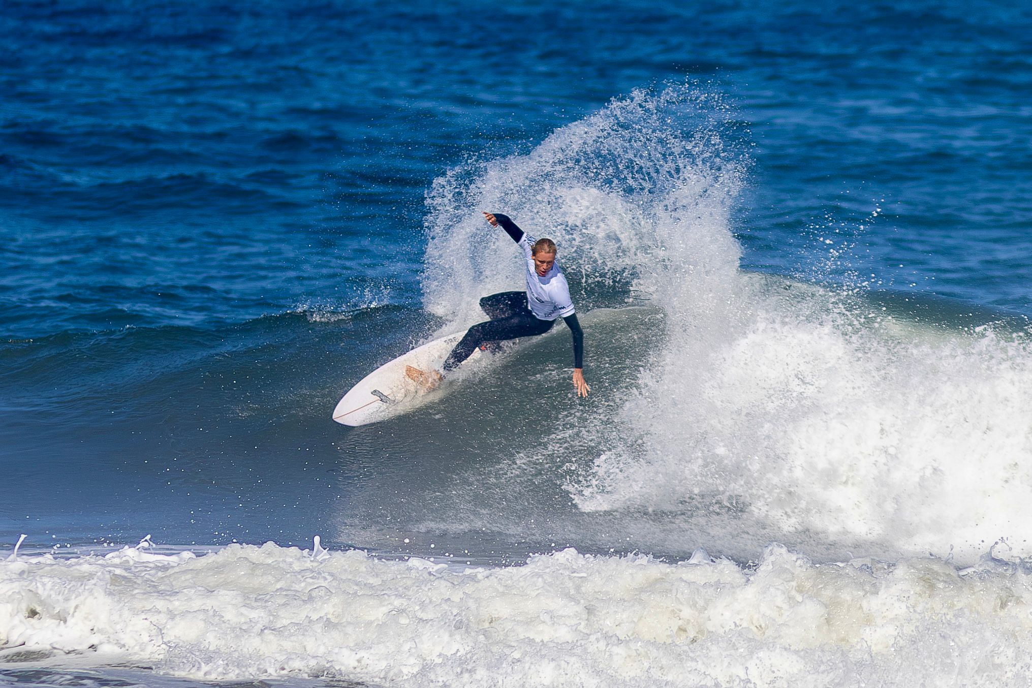 Woman surfer in a white competition jersey carves across a dark blue wave in Leça da Palmeira